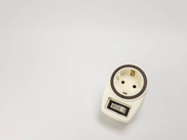 Bu görüntü, düz beyaz bir arkaplan üzerine kurulmuş entegre bir açma kapama düğmesi olan bir elektrik prizinin yakın çekim görüntüsünü içerir. Priz bir krem rengi ve düğme siyah beyaz. Kompozisyon basit, nesneyi vurguluyor. Th