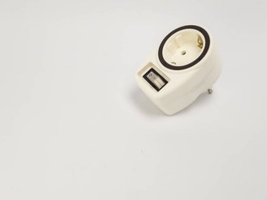 Beyaz bir elektrik adaptörünün yakın plan görüntüsü. Temiz beyaz bir arkaplanda köşegen üzerine kurulmuş bir açma kapama düğmesi var. Adaptörde tek bir soket ve elektrik enerjisi için tasarlanmış standart bir fiş var. Keskin bir odak noktası var. 
