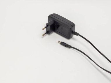 Siyah bir güç adaptörünün yakın çekim görüntüsü. Çift eksenli prizi ve DC konnektörüyle biten siyah bir kablosu var. Adaptör, formunu ve işlevini vurgulayarak, temiz beyaz bir arkaplana karşı izole edilir. Görüntü iyi aydınlatılmış, gösterişli detaylar.