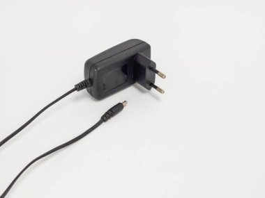Siyah bir güç adaptörünün iki eksenli fişi ile yakın plan bir stüdyo çekimi. Adaptör temiz beyaz bir arkaplanda konumlandırılmış. Fiş, kablo ve adaptörün kaplamasının detaylarını vurguluyor. Görüntü bir simp hissi iletir