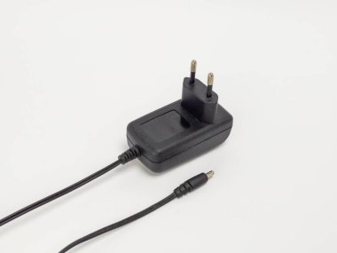 Beyaz bir yüzey üzerinde siyah bir güç adaptörünün kablo ve küçük yuvarlak bir konnektörüyle yakın çekim görüntüsü. Adaptör çerçevenin merkezindedir. Kablo adaptörden uzanıyor. Kompozisyon basit ve temiz. Işıklandırma yumuşak ve eşit.