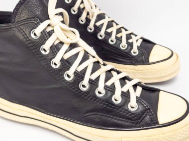 Krem renkli bağcıklı, siyah, yüksek topuklu spor ayakkabıların detaylı bir çekimi. Ayakkabılar beyaz arka plana yerleştiriliyor. Derinin dokusuna, bağcıkların tasarımına ve ayakkabının genel estetiğine odaklanıyor.. 