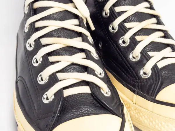 Krem renkli bağcıklı, siyah, yüksek topuklu spor ayakkabıların detaylı bir çekimi. Ayakkabılar beyaz arka plana yerleştiriliyor. Derinin dokusuna, bağcıkların tasarımına ve ayakkabının genel estetiğine odaklanıyor.. 