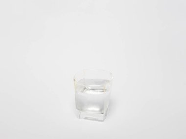 Temiz suyla dolu bir bardağın yakın plan çekimi, düz beyaz arka plan üzerine kurulmuş. Cam basit ve modern tasarım, kare tabanlı ve yuvarlak. Su berrak ve ferahlatıcı görünüyor..