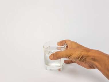 Bir bardak suya uzanan bir elin yakın plan çekimi. Bardak kısmen temiz suyla doludur ve arka plan düz beyazdır. Görüntü hidratlaşmayı, sağlığı ve içki içme eylemini vurgular..