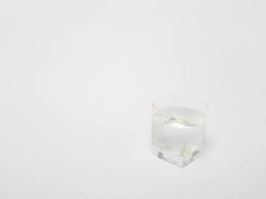 Temiz suyla dolu bir bardağın yakın plan çekimi, düz beyaz arka plan üzerine kurulmuş. Cam basit ve modern tasarım, kare tabanlı ve yuvarlak. Su berrak ve ferahlatıcı görünüyor..