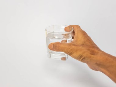 Bir bardak suya uzanan bir elin yakın plan çekimi. Bardak kısmen temiz suyla doludur ve arka plan düz beyazdır. Görüntü hidratlaşmayı, sağlığı ve içki içme eylemini vurgular..