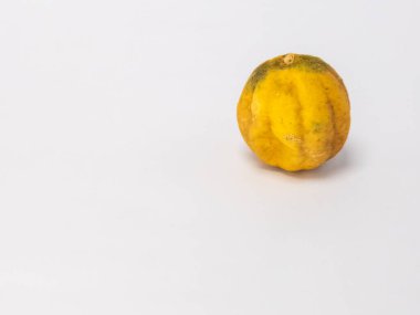 Bu resim, buruşuk, sarı turunçgil meyvesinin, muhtemelen limon ya da benzeri bir meyvenin, temiz, sade beyaz bir arka planda yakın plan görüntüsünü içerir. Meyvenin dokusu ve rengi eski ya da biraz çürümüş olabileceğini gösteriyor..
