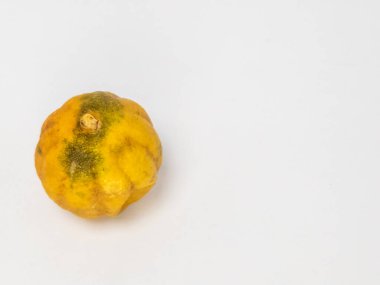 Bu resim, buruşuk, sarı turunçgil meyvesinin, muhtemelen limon ya da benzeri bir meyvenin, temiz, sade beyaz bir arka planda yakın plan görüntüsünü içerir. Meyvenin dokusu ve rengi eski ya da biraz çürümüş olabileceğini gösteriyor..