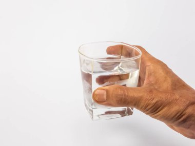 Bir bardak suya uzanan bir elin yakın plan çekimi. Bardak kısmen temiz suyla doludur ve arka plan düz beyazdır. Görüntü hidratlaşmayı, sağlığı ve içki içme eylemini vurgular..