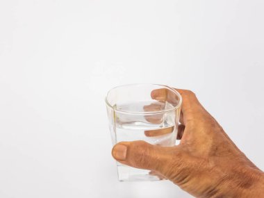 Bir bardak suya uzanan bir elin yakın plan çekimi. Bardak kısmen temiz suyla doludur ve arka plan düz beyazdır. Görüntü hidratlaşmayı, sağlığı ve içki içme eylemini vurgular..