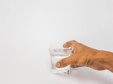 Bir bardak suya uzanan bir elin yakın plan çekimi. Bardak kısmen temiz suyla doludur ve arka plan düz beyazdır. Görüntü hidratlaşmayı, sağlığı ve içki içme eylemini vurgular..