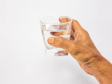 Bir bardak suya uzanan bir elin yakın plan çekimi. Bardak kısmen temiz suyla doludur ve arka plan düz beyazdır. Görüntü hidratlaşmayı, sağlığı ve içki içme eylemini vurgular..