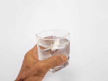 Bir bardak suya uzanan bir elin yakın plan çekimi. Bardak kısmen temiz suyla doludur ve arka plan düz beyazdır. Görüntü hidratlaşmayı, sağlığı ve içki içme eylemini vurgular..