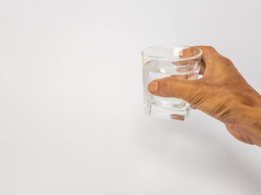 Bir bardak suya uzanan bir elin yakın plan çekimi. Bardak kısmen temiz suyla doludur ve arka plan düz beyazdır. Görüntü hidratlaşmayı, sağlığı ve içki içme eylemini vurgular..