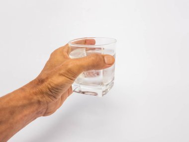 Bir bardak suya uzanan bir elin yakın plan çekimi. Bardak kısmen temiz suyla doludur ve arka plan düz beyazdır. Görüntü hidratlaşmayı, sağlığı ve içki içme eylemini vurgular..