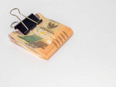 Siyah bir klasör klibiyle tutturulmuş Endonezya Rupiah banknotlarının yer aldığı yakın plan bir fotoğraf. Banknotlar odaklı ve arka plan bembeyaz. Görüntü para, tasarruf ve para gibi finansal kavramları iletir.