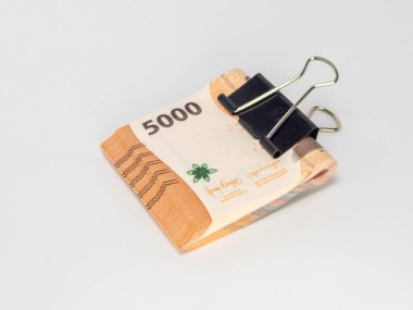 Siyah bir klasör klibiyle tutturulmuş Endonezya Rupiah banknotlarının yer aldığı yakın plan bir fotoğraf. Banknotlar odaklı ve arka plan bembeyaz. Görüntü para, tasarruf ve para gibi finansal kavramları iletir.