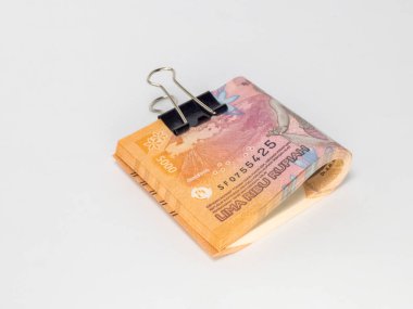 Siyah bir klasör klibiyle tutturulmuş Endonezya Rupiah banknotlarının yer aldığı yakın plan bir fotoğraf. Banknotlar odaklı ve arka plan bembeyaz. Görüntü para, tasarruf ve para gibi finansal kavramları iletir.