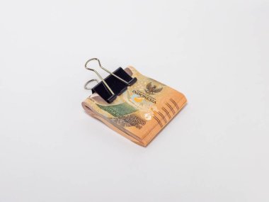 Siyah bir klasör klibiyle tutturulmuş Endonezya Rupiah banknotlarının yer aldığı yakın plan bir fotoğraf. Banknotlar odaklı ve arka plan bembeyaz. Görüntü para, tasarruf ve para gibi finansal kavramları iletir.