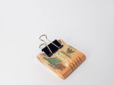 Siyah bir klasör klibiyle tutturulmuş Endonezya Rupiah banknotlarının yer aldığı yakın plan bir fotoğraf. Banknotlar odaklı ve arka plan bembeyaz. Görüntü para, tasarruf ve para gibi finansal kavramları iletir.