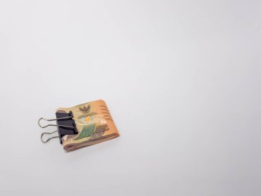 Siyah bir klasör klibiyle tutturulmuş Endonezya Rupiah banknotlarının yer aldığı yakın plan bir fotoğraf. Banknotlar odaklı ve arka plan bembeyaz. Görüntü para, tasarruf ve para gibi finansal kavramları iletir.