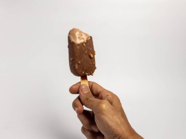 Kısmen yenmiş çikolatalı dondurmayı fındıklı tutarken çekilmiş yakın plan bir fotoğraf. Dondurma hafifçe eriyor ve arka plan bembeyaz. Görüntüde basit bir kompozisyon var, ana konu olarak dondurmaya odaklanıyor..