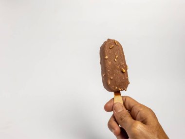 Çikolata kaplı bir dondurmayı tahta bir sopayla sade beyaz arka planda tutarken çekilmiş yakın plan bir fotoğraf. Dondurmanın zengin, kahverengi bir rengi var. Çikolata aromalı ve küçük fındık parçalarıyla kaplı..