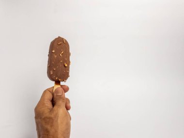 Çikolata kaplı bir dondurmayı tahta bir sopayla sade beyaz arka planda tutarken çekilmiş yakın plan bir fotoğraf. Dondurmanın zengin, kahverengi bir rengi var. Çikolata aromalı ve küçük fındık parçalarıyla kaplı..