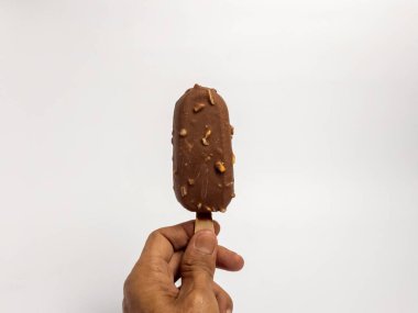 Çikolata kaplı bir dondurmayı tahta bir sopayla sade beyaz arka planda tutarken çekilmiş yakın plan bir fotoğraf. Dondurmanın zengin, kahverengi bir rengi var. Çikolata aromalı ve küçük fındık parçalarıyla kaplı..