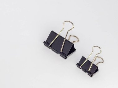 Bu resim, temiz beyaz arkaplana karşı hafif bir açıyla yerleştirilmiş iki siyah klasör klipsi içerir. Ana odak noktaları kliplerdir, tasarımlarını ve metal saplarının etkileşimini gösterirler..