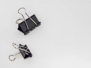 Bu resim, temiz beyaz arkaplana karşı hafif bir açıyla yerleştirilmiş iki siyah klasör klipsi içerir. Ana odak noktaları kliplerdir, tasarımlarını ve metal saplarının etkileşimini gösterirler..