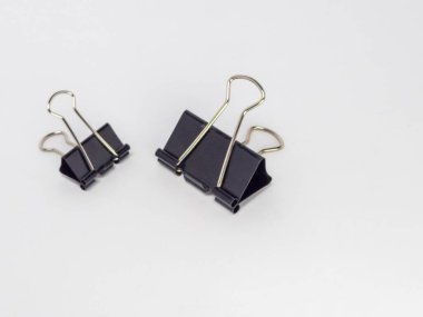 Bu resim, temiz beyaz arkaplana karşı hafif bir açıyla yerleştirilmiş iki siyah klasör klipsi içerir. Ana odak noktaları kliplerdir, tasarımlarını ve metal saplarının etkileşimini gösterirler..