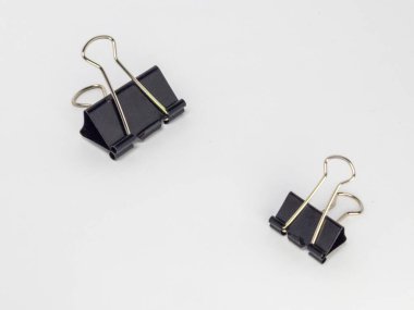 Bu resim, temiz beyaz arkaplana karşı hafif bir açıyla yerleştirilmiş iki siyah klasör klipsi içerir. Ana odak noktaları kliplerdir, tasarımlarını ve metal saplarının etkileşimini gösterirler..