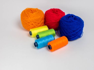 Kırmızı, mavi, sarı, yeşil ve turuncu da dahil olmak üzere çeşitli iplik makaraları ve topları içeren canlı bir kompozisyon. Renkli makaralar, daha büyük iplik toplarının yanına dizilmiştir, hepsi temiz beyaz arka plana karşıdır..