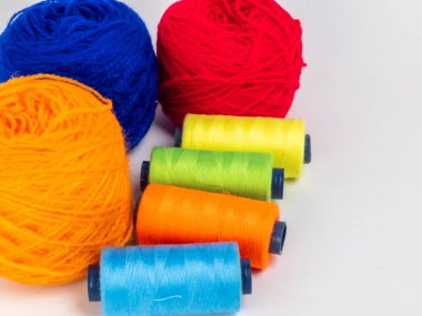 Kırmızı, mavi, sarı, yeşil ve turuncu da dahil olmak üzere çeşitli iplik makaraları ve topları içeren canlı bir kompozisyon. Renkli makaralar, daha büyük iplik toplarının yanına dizilmiştir, hepsi temiz beyaz arka plana karşıdır..