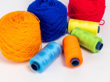 Kırmızı, mavi, sarı, yeşil ve turuncu da dahil olmak üzere çeşitli iplik makaraları ve topları içeren canlı bir kompozisyon. Renkli makaralar, daha büyük iplik toplarının yanına dizilmiştir, hepsi temiz beyaz arka plana karşıdır..