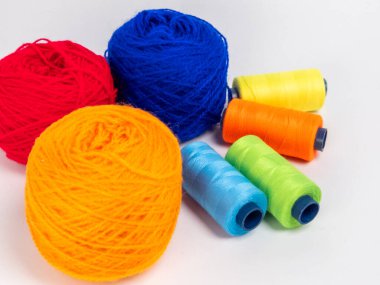 Kırmızı, mavi, sarı, yeşil ve turuncu da dahil olmak üzere çeşitli iplik makaraları ve topları içeren canlı bir kompozisyon. Renkli makaralar, daha büyük iplik toplarının yanına dizilmiştir, hepsi temiz beyaz arka plana karşıdır..