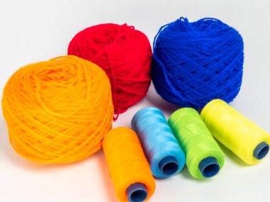 Kırmızı, mavi, sarı, yeşil ve turuncu da dahil olmak üzere çeşitli iplik makaraları ve topları içeren canlı bir kompozisyon. Renkli makaralar, daha büyük iplik toplarının yanına dizilmiştir, hepsi temiz beyaz arka plana karşıdır..