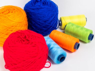 Kırmızı, mavi, sarı, yeşil ve turuncu da dahil olmak üzere çeşitli iplik makaraları ve topları içeren canlı bir kompozisyon. Renkli makaralar, daha büyük iplik toplarının yanına dizilmiştir, hepsi temiz beyaz arka plana karşıdır..