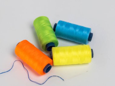 Dört renkli iplik makarasından oluşan yeşil, turuncu, sarı ve mavi renkli bir kolleksiyon, yanında beyaz arkaplanda mavi iplikli bir iğne. Kompozisyon basit ve temiz, makaralar görsel olarak çekici bir şekilde düzenlenmiş..