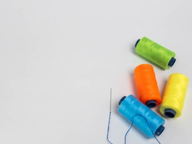 Dört renkli iplik makarasından oluşan yeşil, turuncu, sarı ve mavi renkli bir kolleksiyon, yanında beyaz arkaplanda mavi iplikli bir iğne. Kompozisyon basit ve temiz, makaralar görsel olarak çekici bir şekilde düzenlenmiş..