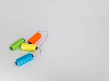 Dört renkli iplik makarasından oluşan yeşil, turuncu, sarı ve mavi renkli bir kolleksiyon, yanında beyaz arkaplanda mavi iplikli bir iğne. Kompozisyon basit ve temiz, makaralar görsel olarak çekici bir şekilde düzenlenmiş..