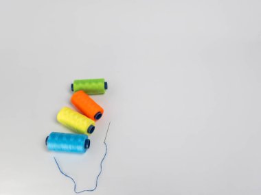 Dört renkli iplik makarasından oluşan yeşil, turuncu, sarı ve mavi renkli bir kolleksiyon, yanında beyaz arkaplanda mavi iplikli bir iğne. Kompozisyon basit ve temiz, makaralar görsel olarak çekici bir şekilde düzenlenmiş..