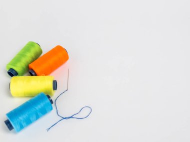 Dört renkli iplik makarasından oluşan yeşil, turuncu, sarı ve mavi renkli bir kolleksiyon, yanında beyaz arkaplanda mavi iplikli bir iğne. Kompozisyon basit ve temiz, makaralar görsel olarak çekici bir şekilde düzenlenmiş..