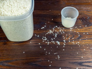 Yakın plan çekimde bir kap pirinç ve içinde biraz pirinç bulunan bir ölçü kabı ve koyu ahşap bir yüzeyde dağınık pirinç taneleri görülüyor. Resim, yemek hazırlama, yemek pişirme ve malzemelere vurgu yapılan bir mutfak sahnesini çekiyor..