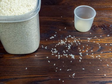 Tepeden tırnağa bir çekimde ölçü kabının yanında beyaz pirinç konteynırı sergilenir. Dağıtılmış pirinç taneleri zengin kahverengi ahşap yüzey.