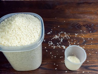 Temiz bir kabın içinde, koyu ahşap bir yüzeye saçılmış tanecikli beyaz pirincin yakın plan çekimi. Pirinçle dolu bir ölçü kabı yakınlarda duruyor..