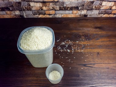 Yakın plan çekimde beyaz pirinçle dolup taşan büyük bir konteynır görülüyor. Yanında aynı pirinçle dolu küçük bir ölçü kabı var. Sahne koyu, ahşap bir yüzeye kurulmuş, dağınık taneciklerle, tuğladan yapılmış bir arka plana karşı..