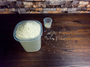 Yakından çekilen bir fotoğrafta, koyu ahşap bir yüzeye dağılmış pirinç taneleriyle birlikte ölçüm kabının yanında içi beyaz pirinçle dolu bir kap sergileniyor. Bu resim yemek pişirme, yemek hazırlama ve mutfak temalarını vurgulamak için hazırlanan malzemeleri tasvir eder..